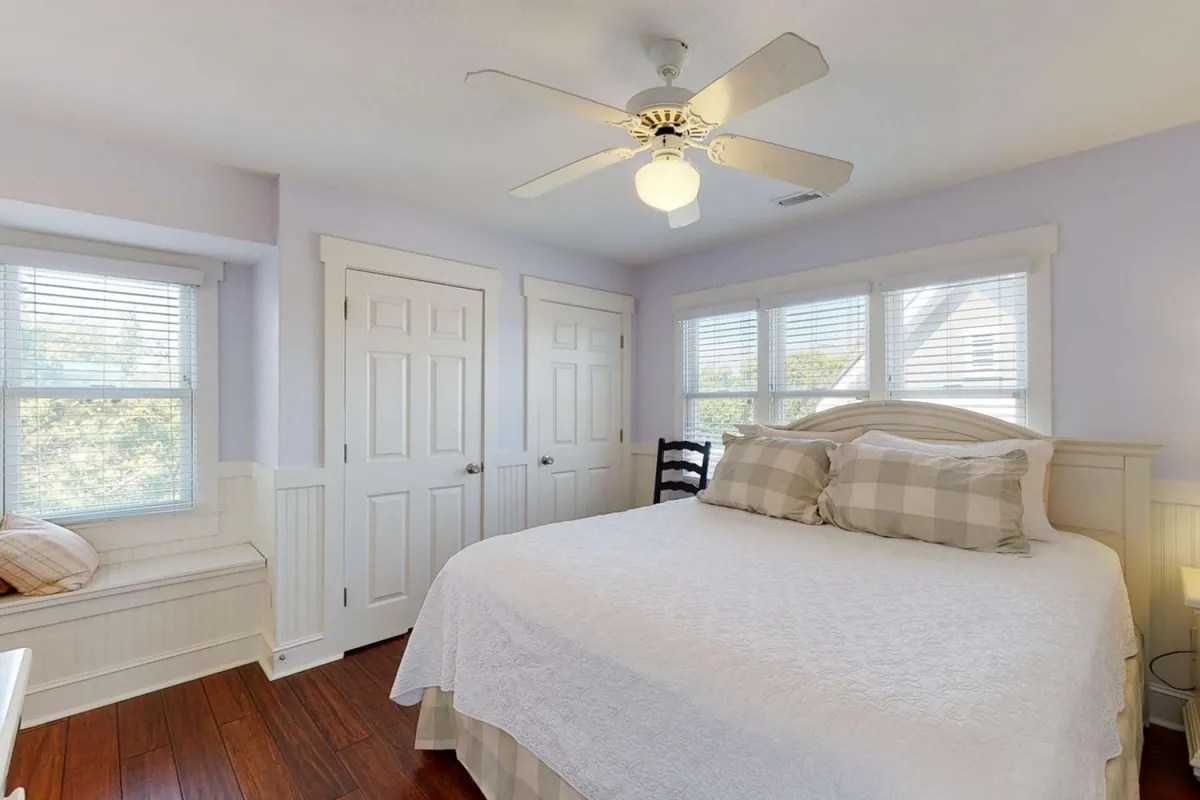 A sun-filled bedroom featuring a comfortable bed with crisp white bedding. Large windows let in natural light and offer tranquil views, while a ceiling fan adds comfort. The room exudes a calming, inviting vibe, perfect for restful nights or quiet reading time.