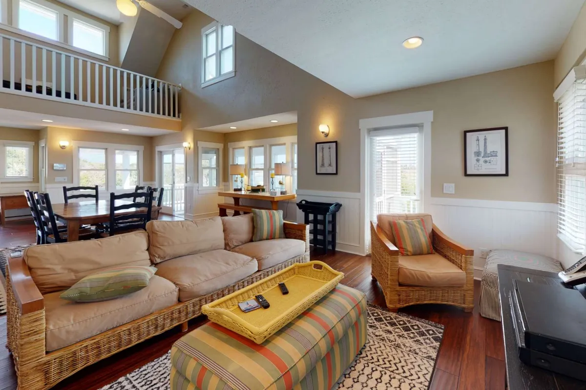 A comfortable living room scene featuring a plush sofa, striped ottoman, dining area, and large windows that let in abundant sunshine. The inviting layout is designed for both relaxation and socializing, with easy access between seating and dining spaces.