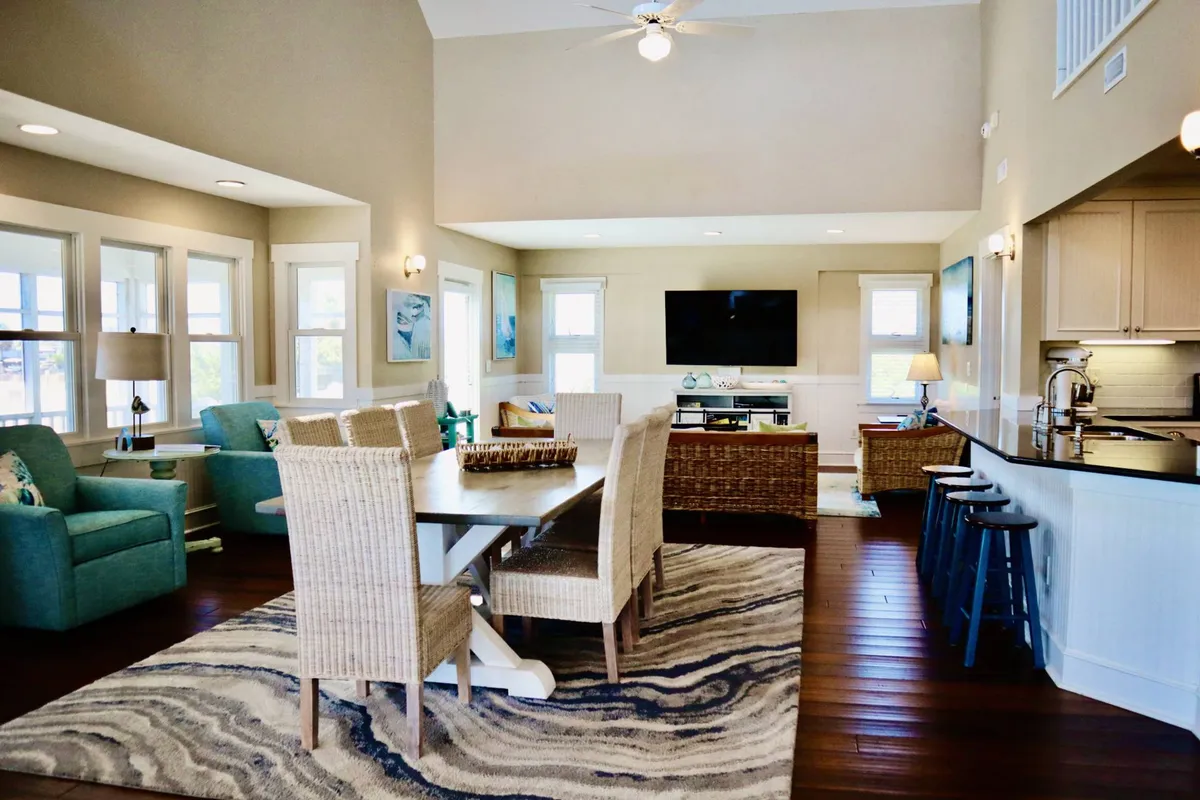 A spacious, sunlit open-plan living area featuring a cozy sofa seating arrangement, a large television for movie nights, a welcoming dining table, and a modern kitchen island all in view. Large windows and vaulted ceilings allow natural light to flood the space, creating a warm, inviting atmosphere ideal for gatherings and relaxation.