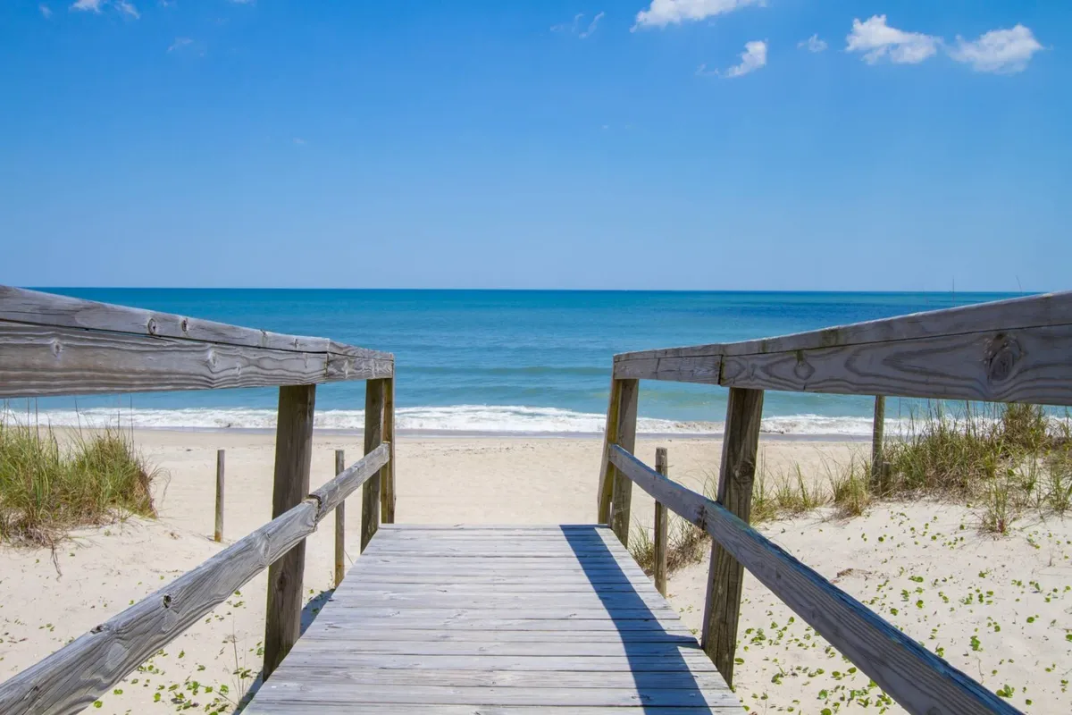 A wide wooden walkway leads directly onto a sandy beach, with gentle ocean waves rolling in beneath a bright blue sky. The area is open, inviting, and perfect for leisurely walks or letting dogs explore. The natural dunes and clear shoreline provide a quintessential island experience.
