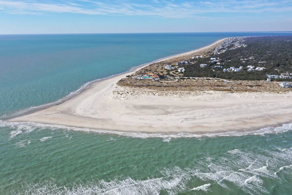 A wide, inviting beach scene with gentle ocean waves meeting the sandy shore. In the distance, residential areas are visible, suggesting easy access from home to beach. The beach is ideal for walking, swimming, and exploring tidal pools, making it perfect for multi-generational family adventures.