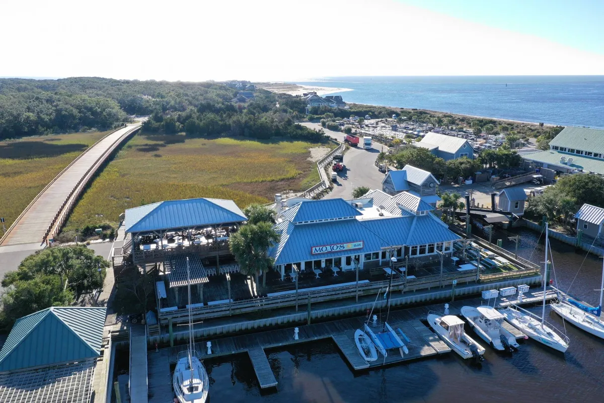 A relaxed restaurant deck sits above the marina, with boats and greenery creating a coastal ambiance. The deck is set up for outdoor dining with expansive views over the water, embodying the ease of waterfront meals and the island’s social atmosphere.
